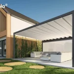 Pergola tarasowa wolnostojąca Tarasola Flat, zainstalowana na przestronnym tarasie z jasnych, kompozytowych desek. Aluminiowa rama w kolorze głębokiego antracytu wspiera białe, tkaninowe zadaszenie rzymskie, które jest rozłożone i układa się w charakterystyczne, miękkie fale podwieszone na poprzecznych profilach. Konstrukcja charakteryzuje się masywną górną belką, która pełni funkcję estetyczną oraz techniczną, skrywając system odprowadzania wody. Pod pergolą zaaranżowano luksusową strefę wypoczynku z białym meblem narożnym i niskim stolikiem kawowym, osłoniętą w pełni rozwiniętym refleksolem Zip. W tle widoczny jest nowoczesny budynek z elewacją łączącą biel, cegłę oraz duże przeszklenia, a także starannie zaprojektowany ogród z trawnikiem i ozdobnymi trawami.