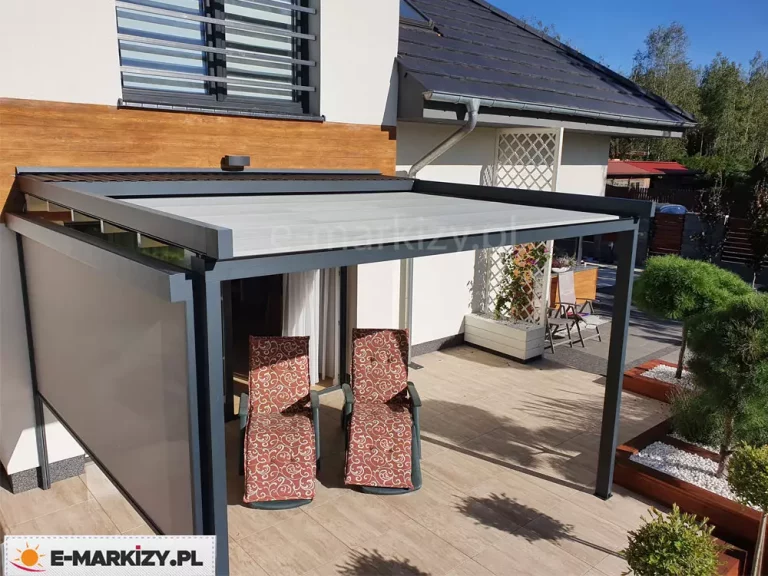 Zdjęcie prezentuje nowoczesną pergolę tarasową Luxo zamontowaną do elewacji budynku, wyposażoną w dodatkową osłonę boczną - refleksol Zip. Konstrukcja aluminiowa w kolorze ciemnego antracytu posiada w pełni rozwinięty dach z jasnej tkaniny oraz opuszczoną roletę pionową typu screen na jednym z boków, która zapewnia dodatkową prywatność i ochronę przed słońcem. Na tarasie wyłożonym jasnobrązowymi płytami imitującymi drewno ustawiono dwa leżaki z wzorzystymi, brązowo-czerwonymi materacami. Elewacja budynku łączy biały tynk z dekoracyjnymi panelami o strukturze drewna. W tle widoczny jest fragment ogrodu z ozdobnymi roślinami w donicach, biała ażurowa kratka oraz sąsiednia zabudowa pod bezchmurnym niebem.