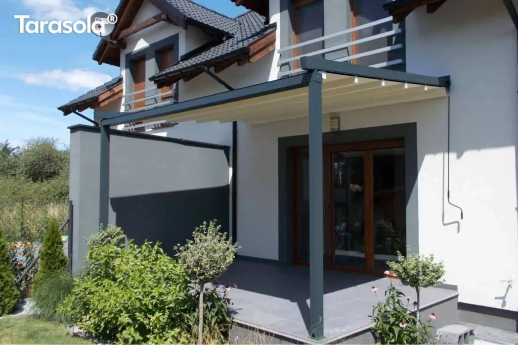 Nowoczesna pergola tarasowa przyścienna Tarasola Simple o konstrukcji przyściennej w kolorze ciemnoszarym (antracytowym), zamontowana nad tarasem wyłożonym dużymi, grafitowymi płytami. Zadaszenie stanowi ruchoma tkanina techniczna w kolorze kremowym, która jest rozwinięta, odsłaniając widok na aluminiowe profile prowadzące. Konstrukcja wspiera się na dwóch solidnych słupach o przekroju kwadratowym, a jedna z bocznych stron została wyposażona w pełny ekran osłonowy w kolorze dopasowanym do stelaża, zapewniający prywatność i ochronę przed wiatrem. Budynek o jasnej elewacji posiada ciemne obramowania wokół okien tarasowych oraz dopasowane kolorystycznie barierki balkonowe na piętrze. Na tarasie oraz w jego bezpośrednim sąsiedztwie widoczne są rośliny ozdobne w donicach i nasadzenia ogrodowe, w tym niskie krzewy i drzewka formowane w kule. Zdjęcie wykonane w pełnym słońcu, podkreślającym kontrast między jasnym materiałem dachu a ciemną konstrukcją pergoli.