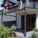 Nowoczesna pergola tarasowa przyścienna Tarasola Simple o konstrukcji przyściennej w kolorze ciemnoszarym (antracytowym), zamontowana nad tarasem wyłożonym dużymi, grafitowymi płytami. Zadaszenie stanowi ruchoma tkanina techniczna w kolorze kremowym, która jest rozwinięta, odsłaniając widok na aluminiowe profile prowadzące. Konstrukcja wspiera się na dwóch solidnych słupach o przekroju kwadratowym, a jedna z bocznych stron została wyposażona w pełny ekran osłonowy w kolorze dopasowanym do stelaża, zapewniający prywatność i ochronę przed wiatrem. Budynek o jasnej elewacji posiada ciemne obramowania wokół okien tarasowych oraz dopasowane kolorystycznie barierki balkonowe na piętrze. Na tarasie oraz w jego bezpośrednim sąsiedztwie widoczne są rośliny ozdobne w donicach i nasadzenia ogrodowe, w tym niskie krzewy i drzewka formowane w kule. Zdjęcie wykonane w pełnym słońcu, podkreślającym kontrast między jasnym materiałem dachu a ciemną konstrukcją pergoli.