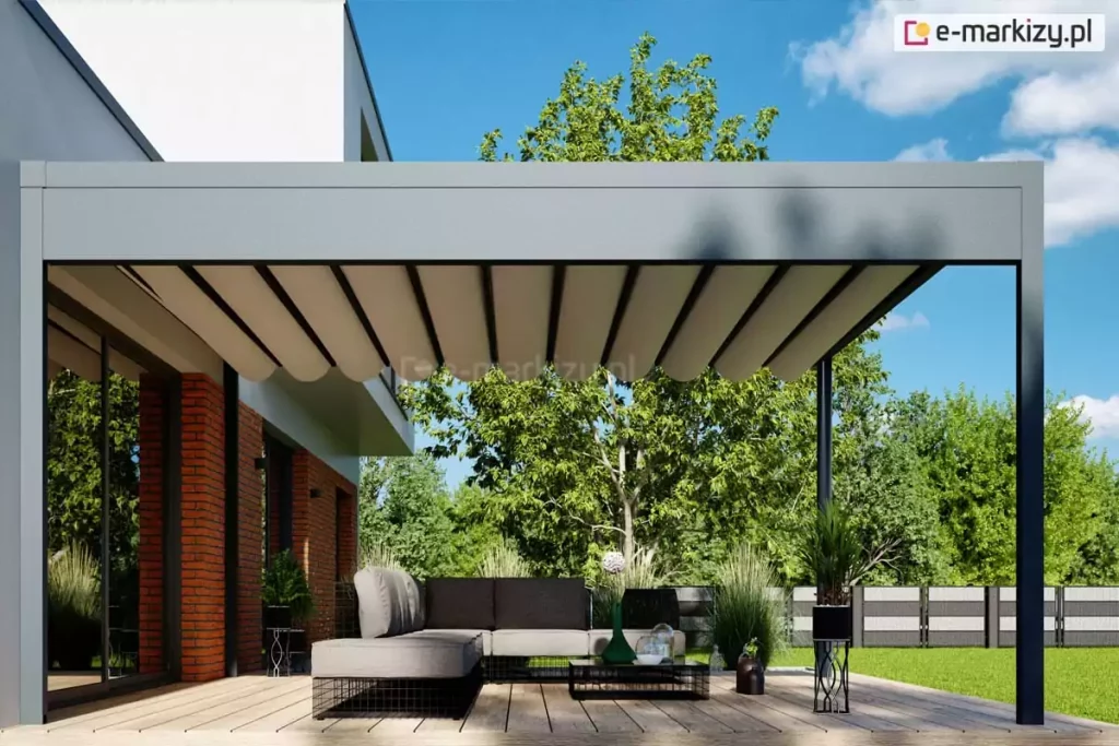 Wizualizacja przedstawia nowoczesną, wolnostojącą pergolę tarasową Luxury o minimalistycznym designie, zamontowaną na przestronnym tarasie z jasnych desek kompozytowych. Konstrukcja pergoli w kolorze matowego antracytu charakteryzuje się szeroką krawędzią dachu, w której ukryty jest system rynnowy do odprowadzania wody opadowej. Pod dachem znajduje się jasne, kremowe poszycie tkaninowe, które jest rozsunięte i układa się w estetyczne, równomierne fale podwieszone na poprzecznych profilach. W strefie wypoczynkowej pod zadaszeniem ustawiono luksusowy, szary narożnik o ażurowej konstrukcji podstawy oraz niski stolik kawowy. Pergola sąsiaduje z nowoczesnym budynkiem o białej elewacji z elementami czerwonej cegły i dużymi przeszkleniami. Tło stanowi zadbany ogród z zielonymi drzewami i trawnikiem, a całość kompozycji dopełniają rośliny doniczkowe w dekoracyjnych stojakach, tworząc spójną i elegancką przestrzeń rekreacyjną w pełnym słońcu.