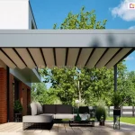 Wizualizacja przedstawia nowoczesną, wolnostojącą pergolę tarasową Luxury o minimalistycznym designie, zamontowaną na przestronnym tarasie z jasnych desek kompozytowych. Konstrukcja pergoli w kolorze matowego antracytu charakteryzuje się szeroką krawędzią dachu, w której ukryty jest system rynnowy do odprowadzania wody opadowej. Pod dachem znajduje się jasne, kremowe poszycie tkaninowe, które jest rozsunięte i układa się w estetyczne, równomierne fale podwieszone na poprzecznych profilach. W strefie wypoczynkowej pod zadaszeniem ustawiono luksusowy, szary narożnik o ażurowej konstrukcji podstawy oraz niski stolik kawowy. Pergola sąsiaduje z nowoczesnym budynkiem o białej elewacji z elementami czerwonej cegły i dużymi przeszkleniami. Tło stanowi zadbany ogród z zielonymi drzewami i trawnikiem, a całość kompozycji dopełniają rośliny doniczkowe w dekoracyjnych stojakach, tworząc spójną i elegancką przestrzeń rekreacyjną w pełnym słońcu.