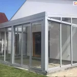 Nowoczesna, biała pergola tarasowa Modes, która została w pełni zabudowana szklanymi panelami, tworząc estetyczny ogród zimowy. Konstrukcja przylega do białej elewacji domu i posiada jednospadowy dach z wyraźnymi profilami. Frontową oraz boczną ścianę tworzą przesuwne, transparentne tafle szkła osadzone w aluminiowych ramach, przy czym boczne przeszklenia pod skosem dachu (tzw. kliny) są zamontowane na stałe. Wewnątrz, na podłożu z jasnych płyt, widoczne są drewniane drzwi prowadzące do budynku, a cała konstrukcja osadzona jest na niskim fundamencie przy zielonym trawniku. W tle, pod bezchmurnym niebem, widać fragment białego pojazdu serwisowego oraz sąsiedni dom z dachem z czerwonej dachówki.