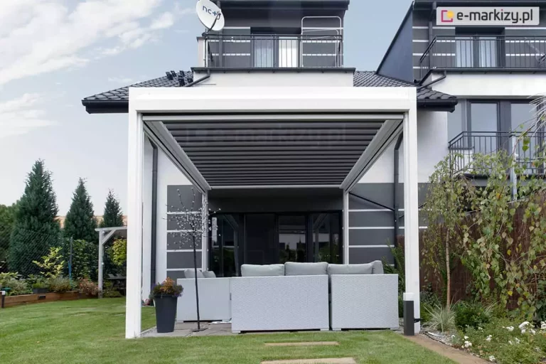 Nowoczesna pergola tarasowa wolnostojąca Sb400 z białą ramą aluminiową i kontrastującym zadaszeniem z lameli w kolorze antracytowym. Pergola została ustawiona na przydomowym tarasie, bezpośrednio przed szklanymi drzwiami przesuwnymi nowoczesnego domu o szaro-białej elewacji. Pod konstrukcją znajduje się duży, narożny zestaw mebli ogrodowych z szarego technorattanu z jasnoszarymi poduszkami. Obok mebli stoi wysoka, czarna donica z roślinami oraz ozdobne drzewko świetlne. W tle widoczny jest zadbany trawnik, gęsty żywopłot z tuj oraz fragment sąsiedniego budynku o identycznej architekturze. Na dachu domu zamontowana jest antena satelitarna. Zdjęcie prezentuje pełną aranżację strefy relaksu, podkreślając dopasowanie kolorystyczne pergoli do detali architektonicznych budynku.