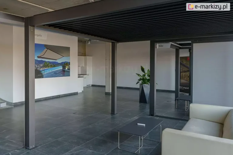 Wnętrze nowoczesnego salonu wystawowego z ekspozycją pergoli aluminiowej Sb350 Aluprof w kolorze antracytowym. Konstrukcja posiada czarny dach z lamelami i wspiera się na kwadratowych słupach zamontowanych do ciemnoszarej, matowej podłogi z wielkoformatowych płyt. Pod pergolą znajduje się strefa wypoczynkowa z kremową sofą oraz niskim, szarym stolikiem kawowym, na którym leży pilot do sterowania. W tle widoczna jest biała ściana z dużym plakatem reklamowym przedstawiającym markizę nad basenem, biała zabudowa meblowa oraz wysoka roślina w nowoczesnej, geometrycznej donicy. Pomieszczenie ma surowy, industrialny sufit z betonu oraz białe ściany, tworząc minimalistyczną przestrzeń ekspozycyjną.
