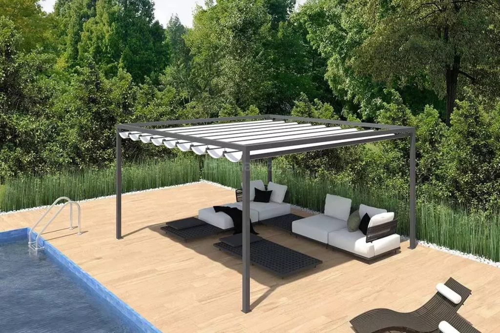 Pergola ogrodowa Dragon wolnostojąca na podeście drewnianym przy basenie zewnętrznym. Pod pergolą w cieniu ustawiony leżak oraz dwie kanapy z białymi poduszkami. Obok na słońcu ustawione dwa leżaki drewniane z białymi poduszkami. Dookoła przestrzeni drewnianego tarasu ogród z wysoką trawą i zielonymi drzewami.