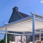 Pergola wolnostojąca Solid Aluprof z podświetlonym dachem tkaninowym ustawiona na tarasie w pobliżu domu wykonanego z kamienia. W budynku, przez duże okna, widoczni ludzie. Nad tarasem widoczne ciemniejące niebo.