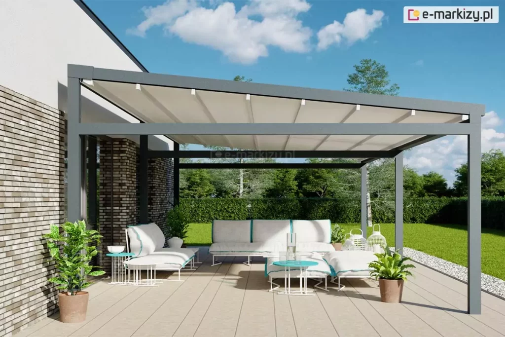 Zdjęcie prezentuje produkt pergola tarasowa wolnostojąca Moderno w aranżacji przyściennej na nowoczesnym tarasie. Konstrukcja pergoli wykonana jest z profili aluminiowych o kwadratowym przekroju w kolorze antracytowym. Zadaszenie stanowi jasna, kremowa tkanina techniczna rozpięta na poprzecznych belkach, zamontowana pod lekkim skosem, co zapewnia swobodny odpływ wody deszczowej. Pergola przylega bezpośrednio do elewacji budynku wykonanej z jasnej cegły klinkierowej z dużymi przeszkleniami. Pod zadaszeniem, na podłodze z jasnych desek kompozytowych, zaaranżowano strefę relaksu z białą sofą modułową ozdobioną błękitnymi lamówkami oraz dopasowanymi pufami i ażurowymi stolikami kawowymi w kolorze białym i turkusowym. Wokół mebli ustawiono rośliny doniczkowe w terakotowych i białych osłonkach oraz dekoracyjne, białe lampiony o różnych kształtach. W tle widoczny jest wypielęgnowany trawnik, zielony żywopłot oraz drzewa liściaste pod błękitnym niebem z rozproszonymi, białymi chmurami. Całość kompozycji jest skąpana w jasnym, naturalnym świetle dziennym, podkreślającym nowoczesną architekturę i detale wykończenia.
