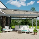 Zdjęcie prezentuje produkt pergola tarasowa wolnostojąca Moderno w aranżacji przyściennej na nowoczesnym tarasie. Konstrukcja pergoli wykonana jest z profili aluminiowych o kwadratowym przekroju w kolorze antracytowym. Zadaszenie stanowi jasna, kremowa tkanina techniczna rozpięta na poprzecznych belkach, zamontowana pod lekkim skosem, co zapewnia swobodny odpływ wody deszczowej. Pergola przylega bezpośrednio do elewacji budynku wykonanej z jasnej cegły klinkierowej z dużymi przeszkleniami. Pod zadaszeniem, na podłodze z jasnych desek kompozytowych, zaaranżowano strefę relaksu z białą sofą modułową ozdobioną błękitnymi lamówkami oraz dopasowanymi pufami i ażurowymi stolikami kawowymi w kolorze białym i turkusowym. Wokół mebli ustawiono rośliny doniczkowe w terakotowych i białych osłonkach oraz dekoracyjne, białe lampiony o różnych kształtach. W tle widoczny jest wypielęgnowany trawnik, zielony żywopłot oraz drzewa liściaste pod błękitnym niebem z rozproszonymi, białymi chmurami. Całość kompozycji jest skąpana w jasnym, naturalnym świetle dziennym, podkreślającym nowoczesną architekturę i detale wykończenia.