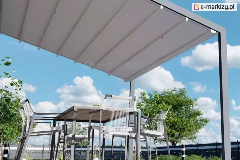 Wizualizacja ujęta z perspektywy żabiej, prezentująca od dołu konstrukcję zadaszenia tarasowego pergola Light tarasola. Na pierwszym planie widoczna jest dolna strona poszycia dachowego wykonanego z jasnej, wysokogatunkowej tkaniny technicznej, która jest rozpięta na poprzecznych profilach przeciwwietrznych. Tkanina tworzy system lekkich, regularnych fałd, charakterystycznych dla rzymskiego systemu składania dachu. Aluminiowy stelaż pergoli w kolorze ciemnego antracytu tworzy geometryczną ramę, a w prawym rogu widoczna jest pionowa kolumna wsporcza o prostokątnym przekroju. Pod zadaszeniem znajduje się strefa jadalniana z nowoczesnym stołem o konstrukcji pasującej do koloru pergoli oraz krzesłami ogrodowymi z charakterystycznymi białymi oparciami z pionowych linek. Perspektywa z dołu eksponuje przestrzeń pod pergolą oraz jej wysokość, otwierając widok na błękitne niebo z licznymi, białymi, puszystymi chmurami typu cumulus. W tle za meblami widać gęstą koronę zielonego drzewa liściastego oraz fragment ciemnego, pasiastego ogrodzenia posesji. Kompozycja zdjęcia podkreśla lekkość wizualną modelu pergola Light tarasola oraz precyzję wykonania detali konstrukcyjnych, takich jak prowadnice i mocowania materiału.