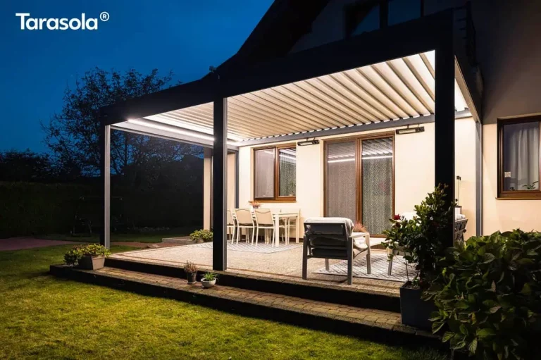 Pergola Essential Lamelowa Przyścienna Galeria Zdjęć przedstawia nowoczesną, bioklimatyczną konstrukcję Essential tarasola w warunkach nocnych, zamontowaną przy jasnej elewacji domu. Pergola w kolorze ciemnego antracytu jest intensywnie oświetlona od wewnątrz za pomocą zintegrowanych listew LED, które emitują jasne, ciepłe światło, oświetlając białe lamele dachowe oraz przestrzeń tarasu. Pod zadaszeniem widoczna jest strefa jadalniana z białym stołem i krzesłami oraz strefa wypoczynkowa z wygodnym fotelem, ustawione na dekoracyjnych dywanach zewnętrznych. Na ścianie budynku, pod dachem pergoli, zamontowane są dwa promienniki ciepła, które zwiększają komfort użytkowania tarasu w chłodniejsze wieczory. Konstrukcja wsparta jest na solidnych słupach, a podłoże tarasu wyłożono jasną kostką brukową z ciemniejszym obramowaniem. W tle, za oświetloną strefą, panuje głęboki mrok nocy, z ledwo widocznymi zarysami drzew i ogrodu, co potęguje kontrast i skupia uwagę na przytulnie urządzonej przestrzeni pod pergolą.