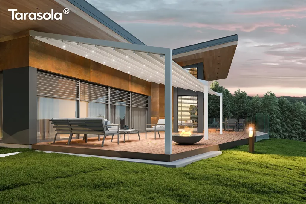 Nowoczesna przyścienna pergola Tarasola Light zamontowana na przestronnym drewnianym tarasie przy nowoczesnym budynku o industrialnej architekturze z elewacją w kolorze rdzawym i szarym. Konstrukcja pergoli wykonana jest z aluminiowych profili w kolorze białym, które podtrzymują rozsuwane zadaszenie tkaninowe o pofalowanej strukturze, również w jasnym odcieniu. Pod materiałowym dachem zintegrowano system oświetlenia LED w postaci punktowych świateł rozmieszczonych równomiernie na poprzecznych profilach, które oświetlają przestrzeń wypoczynkową po zmroku. Na tarasie znajduje się zestaw mebli ogrodowych składający się z szarej sofy z miękkimi poduszkami, stolika kawowego oraz fotela, a w centralnej części umiejscowiono okrągłe palenisko z palącym się ogniem. Tło stanowi przeszklona ściana budynku z zewnętrznymi roletami lamelowymi oraz zielony ogród z przystrzyżonym trawnikiem i gęstymi drzewami iglastymi pod wieczornym, lekko zachmurzonym niebem o fioletowych i różowych odcieniach. Całość kompozycji podkreśla funkcjonalność zadaszenia w aranżacji strefy relaksu, łącząc nowoczesny design z przytulną atmosferą domowego ogrodu.