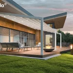 Nowoczesna przyścienna pergola Tarasola Light zamontowana na przestronnym drewnianym tarasie przy nowoczesnym budynku o industrialnej architekturze z elewacją w kolorze rdzawym i szarym. Konstrukcja pergoli wykonana jest z aluminiowych profili w kolorze białym, które podtrzymują rozsuwane zadaszenie tkaninowe o pofalowanej strukturze, również w jasnym odcieniu. Pod materiałowym dachem zintegrowano system oświetlenia LED w postaci punktowych świateł rozmieszczonych równomiernie na poprzecznych profilach, które oświetlają przestrzeń wypoczynkową po zmroku. Na tarasie znajduje się zestaw mebli ogrodowych składający się z szarej sofy z miękkimi poduszkami, stolika kawowego oraz fotela, a w centralnej części umiejscowiono okrągłe palenisko z palącym się ogniem. Tło stanowi przeszklona ściana budynku z zewnętrznymi roletami lamelowymi oraz zielony ogród z przystrzyżonym trawnikiem i gęstymi drzewami iglastymi pod wieczornym, lekko zachmurzonym niebem o fioletowych i różowych odcieniach. Całość kompozycji podkreśla funkcjonalność zadaszenia w aranżacji strefy relaksu, łącząc nowoczesny design z przytulną atmosferą domowego ogrodu.