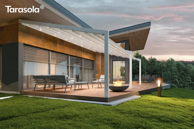 Nowoczesna przyścienna pergola Tarasola Light zamontowana na przestronnym drewnianym tarasie przy nowoczesnym budynku o industrialnej architekturze z elewacją w kolorze rdzawym i szarym. Konstrukcja pergoli wykonana jest z aluminiowych profili w kolorze białym, które podtrzymują rozsuwane zadaszenie tkaninowe o pofalowanej strukturze, również w jasnym odcieniu. Pod materiałowym dachem zintegrowano system oświetlenia LED w postaci punktowych świateł rozmieszczonych równomiernie na poprzecznych profilach, które oświetlają przestrzeń wypoczynkową po zmroku. Na tarasie znajduje się zestaw mebli ogrodowych składający się z szarej sofy z miękkimi poduszkami, stolika kawowego oraz fotela, a w centralnej części umiejscowiono okrągłe palenisko z palącym się ogniem. Tło stanowi przeszklona ściana budynku z zewnętrznymi roletami lamelowymi oraz zielony ogród z przystrzyżonym trawnikiem i gęstymi drzewami iglastymi pod wieczornym, lekko zachmurzonym niebem o fioletowych i różowych odcieniach. Całość kompozycji podkreśla funkcjonalność zadaszenia w aranżacji strefy relaksu, łącząc nowoczesny design z przytulną atmosferą domowego ogrodu.