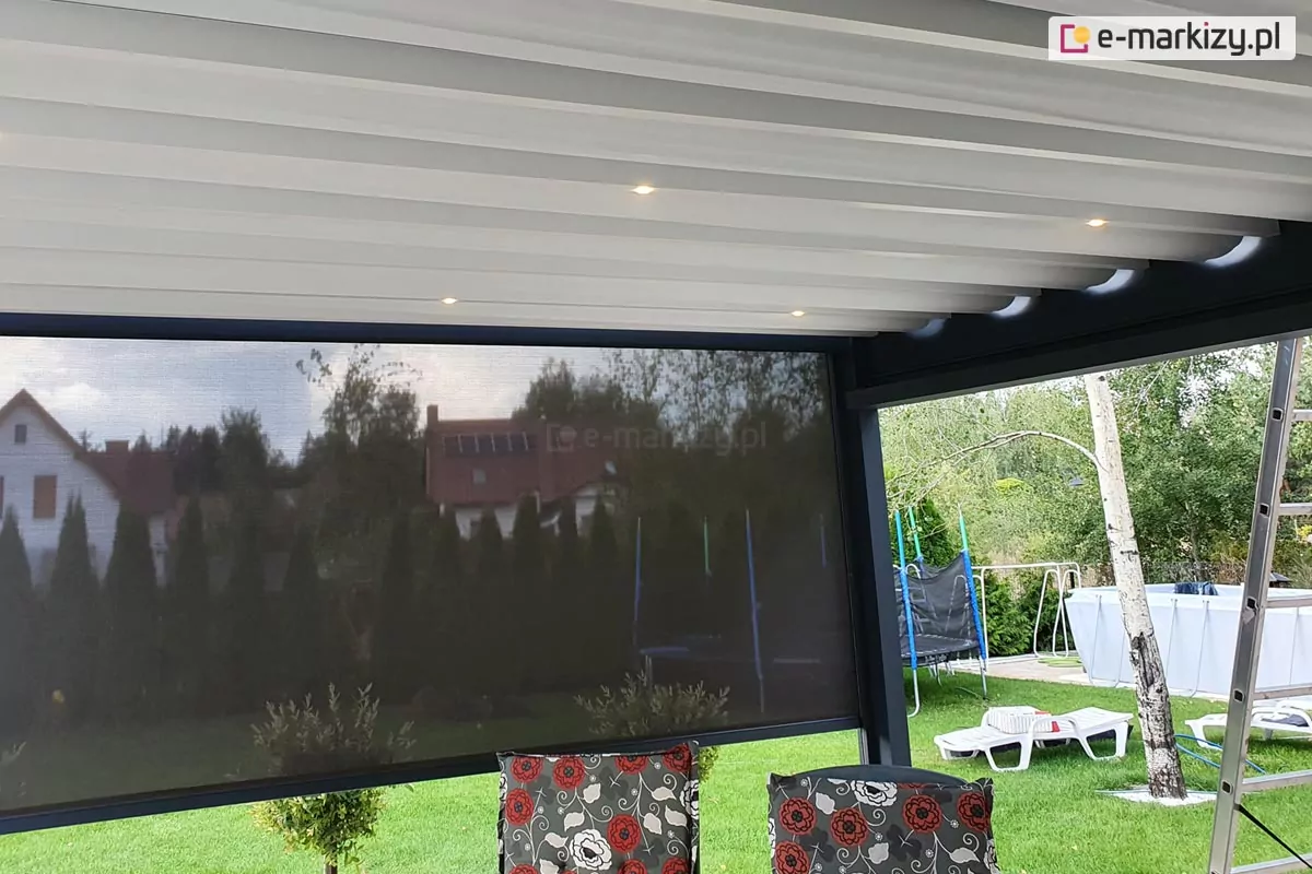 Zdjęcie prezentuje perspektywę od wewnątrz pergoli tarasowej Luxury, ukazującą detale systemu oświetlenia oraz osłon bocznych. Pod jasnym, białym zadaszeniem tkaninowym, które jest rozpięte na poprzecznych profilach, zamontowano zintegrowane punkty świetlne LED emitujące subtelne światło. Na pierwszym planie widoczna jest ciemna, półtransparentna roleta typu screen, która przesłania widok na ogród, redukując nasłonecznienie i zapewniając prywatność. Przez oczka tkaniny screen prześwituje zarys sąsiedniej zabudowy oraz gęsta linia tui. W dolnej części kadru widoczne są oparcia foteli ogrodowych z dekoracyjnymi poduszkami w kwiatowy wzór, a po prawej stronie fragment białej brzozy oraz drabina malarska, co sugeruje etap prac montażowych lub konserwacyjnych przy konstrukcji.