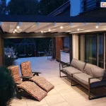 Zdjęcie prezentuje nowoczesną pergolę tarasową Luxury z włączonym oświetleniem LED, wykonane po zmroku. Antracytowa konstrukcja aluminiowa o kubistycznym kształcie posiada jasne, tkaninowe zadaszenie, pod którym na poprzecznych profilach zamontowano punktowe źródła światła o ciepłej barwie. Oświetlenie równomiernie rozjaśnia przestrzeń wypoczynkową na tarasie wyłożonym jasnymi płytami, na którym ustawiono nowoczesną, szarą sofę oraz dwa fotele ogrodowe z wzorzystymi poduszkami w odcieniach brązu. Pergola przylega bezpośrednio do budynku z dużymi przeszkleniami, przez które widać oświetlone wnętrze salonu. W tle widoczny jest ciemny ogród z zarysem drzew oraz plac zabaw, co podkreśla przytulną atmosferę i funkcjonalność systemu zadaszenia jako przedłużenia części mieszkalnej domu.
