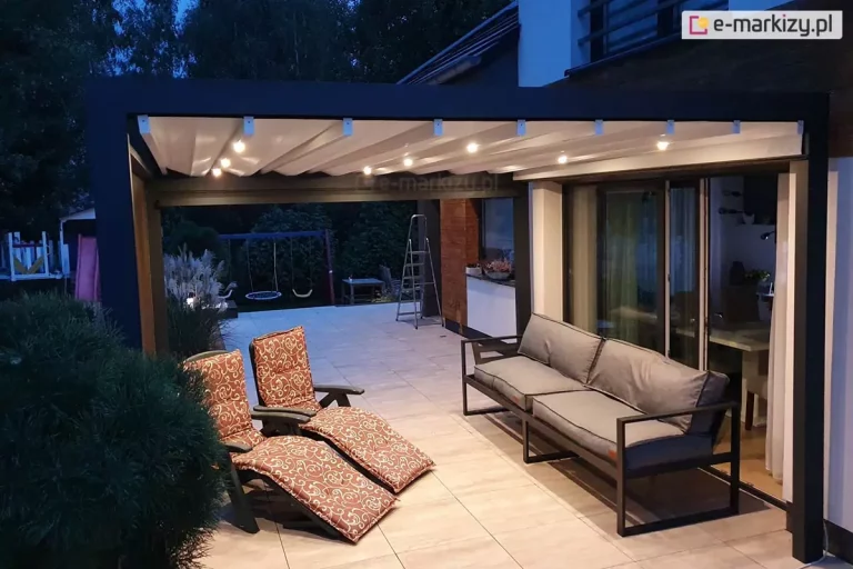 Zdjęcie prezentuje nowoczesną pergolę tarasową Luxury z włączonym oświetleniem LED, wykonane po zmroku. Antracytowa konstrukcja aluminiowa o kubistycznym kształcie posiada jasne, tkaninowe zadaszenie, pod którym na poprzecznych profilach zamontowano punktowe źródła światła o ciepłej barwie. Oświetlenie równomiernie rozjaśnia przestrzeń wypoczynkową na tarasie wyłożonym jasnymi płytami, na którym ustawiono nowoczesną, szarą sofę oraz dwa fotele ogrodowe z wzorzystymi poduszkami w odcieniach brązu. Pergola przylega bezpośrednio do budynku z dużymi przeszkleniami, przez które widać oświetlone wnętrze salonu. W tle widoczny jest ciemny ogród z zarysem drzew oraz plac zabaw, co podkreśla przytulną atmosferę i funkcjonalność systemu zadaszenia jako przedłużenia części mieszkalnej domu.