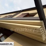 Perspektywa od dołu na złożone zadaszenie tkaninowe nowoczesnej pergoli Luxo w kolorze antracytowym. Kremowy materiał poszycia jest upięty w gęste, estetyczne fałdy pod poziomym profilem aluminiowym, co uwidacznia harmonijkowy system składania tkaniny. Konstrukcja ramy, polakierowana na matowy, ciemny kolor, kontrastuje z jasną elewacją budynku o piaskowym odcieniu. W górnej części kadru widoczna jest brązowa podbitka dachowa oraz fragment systemu rynnowego i ciemnej dachówki, a w tle rozpościera się jasne, lekko zachmurzone niebo. Zdjęcie koncentruje się na detalach mechanizmu zwijania oraz wysokiej jakości wykończenia tekstylnego elementu zadaszenia.