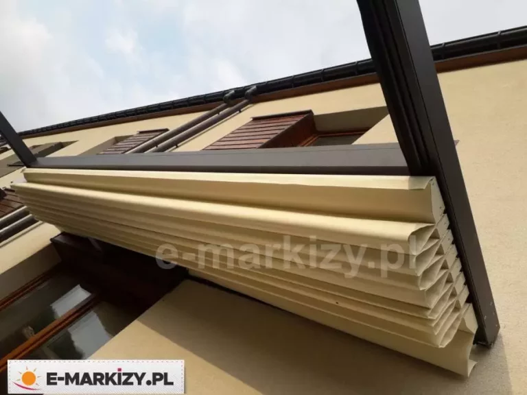 Perspektywa od dołu na złożone zadaszenie tkaninowe nowoczesnej pergoli Luxo w kolorze antracytowym. Kremowy materiał poszycia jest upięty w gęste, estetyczne fałdy pod poziomym profilem aluminiowym, co uwidacznia harmonijkowy system składania tkaniny. Konstrukcja ramy, polakierowana na matowy, ciemny kolor, kontrastuje z jasną elewacją budynku o piaskowym odcieniu. W górnej części kadru widoczna jest brązowa podbitka dachowa oraz fragment systemu rynnowego i ciemnej dachówki, a w tle rozpościera się jasne, lekko zachmurzone niebo. Zdjęcie koncentruje się na detalach mechanizmu zwijania oraz wysokiej jakości wykończenia tekstylnego elementu zadaszenia.