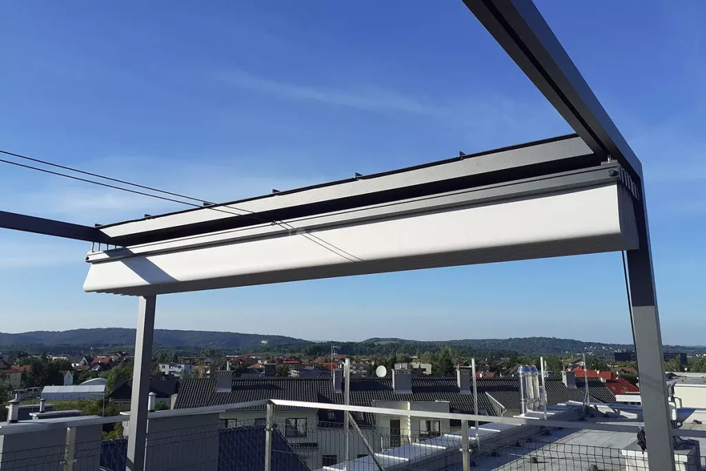 Zbliżenie na mechanizm zadaszenia nowoczesnej pergoli wolnostojącej Dragon o antracytowej, aluminiowej konstrukcji, zamontowanej na tarasie dachowym. Zdjęcie przedstawia jasny, techniczny materiał poszycia, który został całkowicie zwinięty i złożony w pakiety pod górną ramą konstrukcyjną. Widoczna jest precyzyjna praca prowadnic oraz poprzecznych profili, które utrzymują tkaninę w uporządkowanej formie. W górnej części ramy widoczne są również niewielkie czarne wypustki mocujące. Tło stanowi szeroka panorama osiedla domów jednorodzinnych pod czystym, błękitnym niebem. W dolnej części kadru widać fragmenty barierki zabezpieczającej taras oraz kominy wentylacyjne sąsiednich budynków. Perspektywa z dołu podkreśla techniczną estetykę systemu rozsuwanego dachu oraz solidność metalowej ramy.