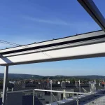Zbliżenie na mechanizm zadaszenia nowoczesnej pergoli wolnostojącej Dragon o antracytowej, aluminiowej konstrukcji, zamontowanej na tarasie dachowym. Zdjęcie przedstawia jasny, techniczny materiał poszycia, który został całkowicie zwinięty i złożony w pakiety pod górną ramą konstrukcyjną. Widoczna jest precyzyjna praca prowadnic oraz poprzecznych profili, które utrzymują tkaninę w uporządkowanej formie. W górnej części ramy widoczne są również niewielkie czarne wypustki mocujące. Tło stanowi szeroka panorama osiedla domów jednorodzinnych pod czystym, błękitnym niebem. W dolnej części kadru widać fragmenty barierki zabezpieczającej taras oraz kominy wentylacyjne sąsiednich budynków. Perspektywa z dołu podkreśla techniczną estetykę systemu rozsuwanego dachu oraz solidność metalowej ramy.