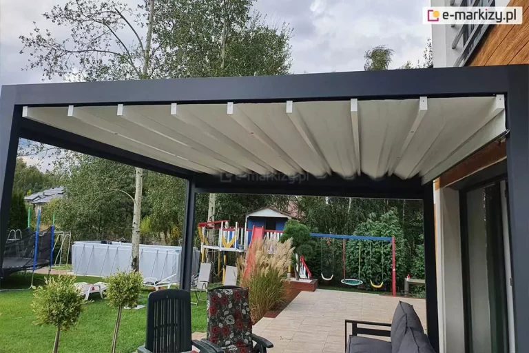Zdjęcie prezentuje przyścienną pergolę tarasową Luxury o nowoczesnej, kubistycznej formie z rozłożonym dachem tkaninowym. Konstrukcja nośna wykonana z grubych profili aluminiowych w kolorze antracytowym tworzy stabilną ramę, pod którą zamontowano system ruchomego zadaszenia. Jasne, białe poszycie dachu jest pofalowane i podwieszone na poprzecznych białych profilach, które przesuwają się po prowadnicach bocznych, umożliwiając regulację stopnia osłonięcia tarasu. Pergola jest zamontowana na wykafelkowanym tarasie przy budynku mieszkalnym z elewacją o strukturze drewna. W tle widoczna jest rekreacyjna część ogrodu z zielonym trawnikiem, drzewami, placem zabaw dla dzieci z huśtawkami oraz basenem stelażowym, co ukazuje praktyczne zastosowanie konstrukcji w przestrzeni przydomowej.