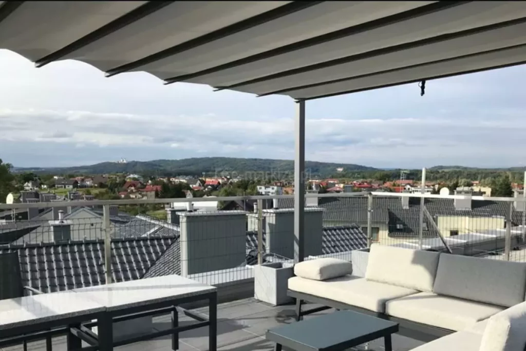 Widok z bliska na zadaszenie tarasowe pergoli wolnostojącej zainstalowanej na dachu budynku, prezentujący system przesuwnego poszycia w kolorze jasnoszarym. Materiał układa się w miękkie, regularne fale podwieszone pod poprzecznymi profilami, które poruszają się wzdłuż bocznych szyn antracytowej ramy. W dolnej części kadru widoczny jest fragment strefy wypoczynkowej z nowoczesną, jasną sofą modułową oraz szarymi stolikami o gładkich blatach. Tło stanowi rozległa panorama podmiejskiej miejscowości z domami o ciemnych dachach i kominach, widoczna zza metalowej balustrady tarasu z wypełnieniem z siatki. Niebo jest jasne, pokryte delikatną warstwą chmur, co nadaje scenie spokojny, zrelaksowany charakter.