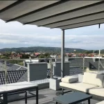 Widok z bliska na zadaszenie tarasowe pergoli wolnostojącej zainstalowanej na dachu budynku, prezentujący system przesuwnego poszycia w kolorze jasnoszarym. Materiał układa się w miękkie, regularne fale podwieszone pod poprzecznymi profilami, które poruszają się wzdłuż bocznych szyn antracytowej ramy. W dolnej części kadru widoczny jest fragment strefy wypoczynkowej z nowoczesną, jasną sofą modułową oraz szarymi stolikami o gładkich blatach. Tło stanowi rozległa panorama podmiejskiej miejscowości z domami o ciemnych dachach i kominach, widoczna zza metalowej balustrady tarasu z wypełnieniem z siatki. Niebo jest jasne, pokryte delikatną warstwą chmur, co nadaje scenie spokojny, zrelaksowany charakter.