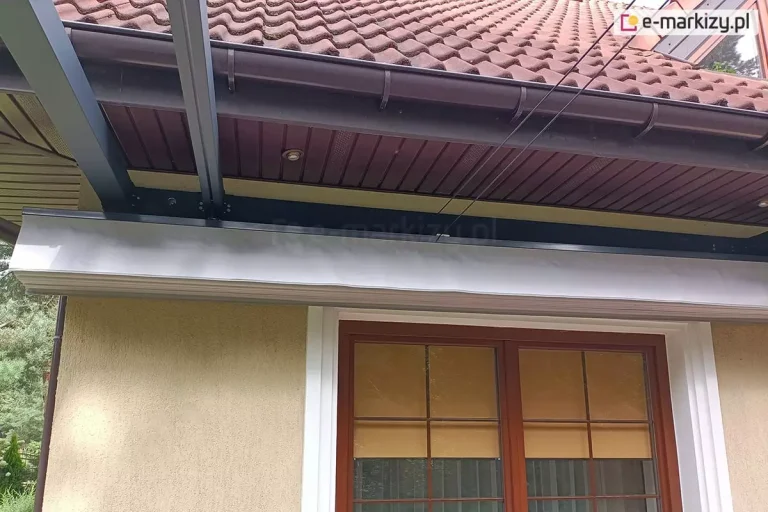 Szerokie ujęcie prezentujące montaż przyścienny pergoli rzymskiej na elewacji domu jednorodzinnego. Aluminiowa konstrukcja w kolorze antracytowym została zamocowana bezpośrednio pod okapem dachu, który wykończony jest brązową podbitką z punktowym oświetleniem LED oraz rynną w tym samym kolorze. Na ścianie budynku, nad brązowym oknem z widocznymi wewnętrznymi roletami materiałowymi, zamontowana jest długa, pozioma belka nośna z systemem prowadnic. Pod belką znajduje się jasnoszare poszycie tkaninowe, które jest obecnie w stanie częściowo zwiniętym, tworząc zwartą strukturę materiału. Od konstrukcji odchodzą w stronę ogrodu dwie prostopadłe belki prowadzące. Ponad systemem zadaszenia widoczne są napięte linki stalowe mechanizmu naciągowego. W tle, po lewej stronie, widoczny jest fragment ogrodu z zielonymi drzewami i krzewami. Zdjęcie doskonale ilustruje integrację nowoczesnej osłony tarasowej z architekturą tradycyjnego budynku krytego dachówką ceramiczną.