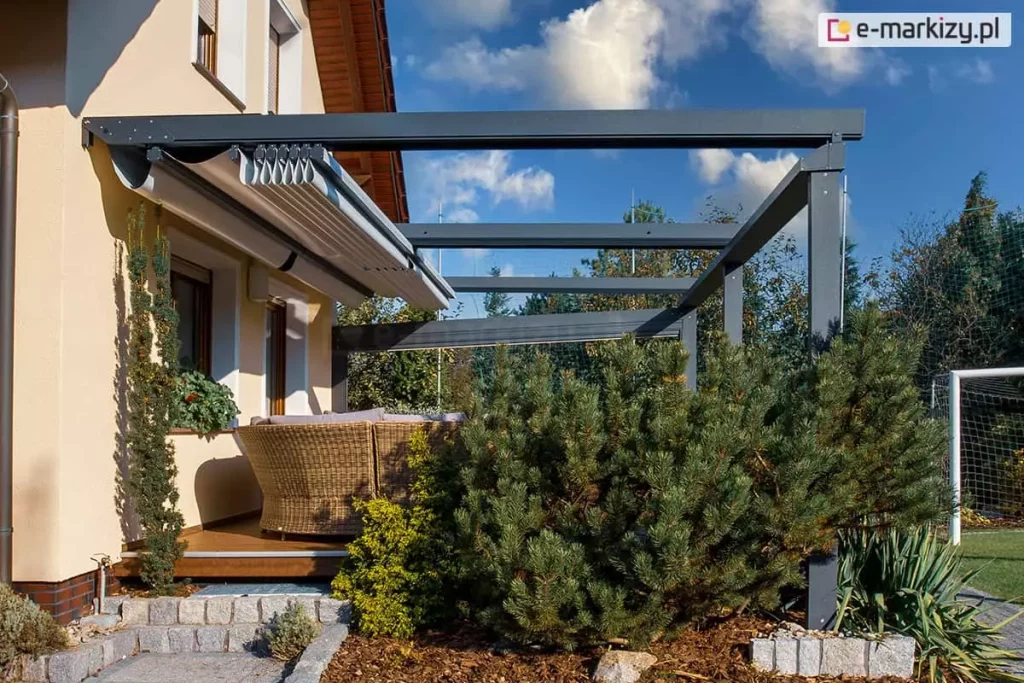 aluminiowa pergola tarasowa w kolorze antracytowym, zainstalowana jako konstrukcja przyścienna bezpośrednio przy elewacji budynku o ciepłej, kremowej kolorystyce. System zadaszenia charakteryzuje się zastosowaniem ruchomej tkaniny technicznej, która na tym ujęciu jest niemal w pełni zsunięta w stronę ściany, co odsłania widok na niebo i wpuszcza więcej światła dziennego do wnętrza tarasu. Mechanizm przesuwny dachu opiera się na szeregu prowadnic oraz poprzecznych profili, które podtrzymują materiał i umożliwiają jego płynne rozkładanie. Konstrukcja wsparta jest na solidnych, pionowych słupach o przekroju kwadratowym, które nadają całości nowoczesnego, industrialnego charakteru. Taras wyłożony jest drewnianą deską, na której ustawiono wypoczynkowe meble z technorattanu. W bezpośrednim otoczeniu pergoli znajduje się starannie zaprojektowany ogród, w którym dominują iglaki o zróżnicowanej wielkości, niskie krzewy ozdobne oraz wyściółka z kory, a granica strefy wypoczynkowej została wyraźnie zaznaczona obrzeżami z jasnej kostki brukowej. W dalszym tle, poza konstrukcją pergoli, dostrzegalny jest fragment ogrodzenia oraz siatka bramki piłkarskiej, co sugeruje lokalizację w przydomowej strefie rekreacyjnej. Całość kompozycji wizualnej dokumentuje estetyczne połączenie nowoczesnych rozwiązań architektonicznych z naturalnym otoczeniem przydomowym, gdzie metalowa konstrukcja pergoli stanowi kontrastowy, lecz spójny element dla jasnej elewacji domu.