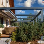 aluminiowa pergola tarasowa w kolorze antracytowym, zainstalowana jako konstrukcja przyścienna bezpośrednio przy elewacji budynku o ciepłej, kremowej kolorystyce. System zadaszenia charakteryzuje się zastosowaniem ruchomej tkaniny technicznej, która na tym ujęciu jest niemal w pełni zsunięta w stronę ściany, co odsłania widok na niebo i wpuszcza więcej światła dziennego do wnętrza tarasu. Mechanizm przesuwny dachu opiera się na szeregu prowadnic oraz poprzecznych profili, które podtrzymują materiał i umożliwiają jego płynne rozkładanie. Konstrukcja wsparta jest na solidnych, pionowych słupach o przekroju kwadratowym, które nadają całości nowoczesnego, industrialnego charakteru. Taras wyłożony jest drewnianą deską, na której ustawiono wypoczynkowe meble z technorattanu. W bezpośrednim otoczeniu pergoli znajduje się starannie zaprojektowany ogród, w którym dominują iglaki o zróżnicowanej wielkości, niskie krzewy ozdobne oraz wyściółka z kory, a granica strefy wypoczynkowej została wyraźnie zaznaczona obrzeżami z jasnej kostki brukowej. W dalszym tle, poza konstrukcją pergoli, dostrzegalny jest fragment ogrodzenia oraz siatka bramki piłkarskiej, co sugeruje lokalizację w przydomowej strefie rekreacyjnej. Całość kompozycji wizualnej dokumentuje estetyczne połączenie nowoczesnych rozwiązań architektonicznych z naturalnym otoczeniem przydomowym, gdzie metalowa konstrukcja pergoli stanowi kontrastowy, lecz spójny element dla jasnej elewacji domu.