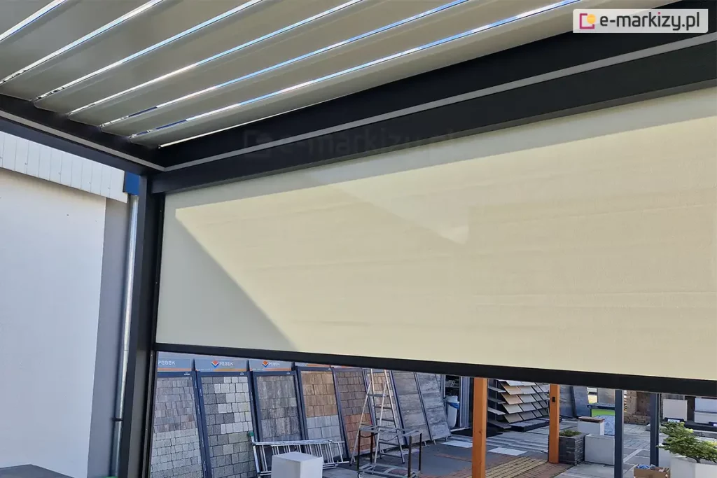 Widok z wnętrza nowoczesnej pergoli tarasowej SB400 na system bocznych osłon przeciwsłonecznych typu screen. Na pierwszym planie widoczny jest refleksol Ziiip 120 Aluprof w kolorze beżowym, który rozwinięty jest na część wysokości, stanowiąc skuteczną barierę przed słońcem i wiatrem. Tkanina charakteryzuje się gładkim splotem i jest stabilnie zamontowana w antracytowej ramie konstrukcyjnej pergoli. Powyżej rolety widoczne jest zadaszenie pergoli wykonane z ruchomych, aluminiowych lameli (system bioklimatyczny) w kolorze białym, ustawionych pod kątem, co pozwala na regulację dopływu światła. W tle, za konstrukcją pergoli, znajduje się ekspozycja zewnętrzna z różnymi rodzajami kostki brukowej, płyt tarasowych oraz elementami małej architektury ogrodowej. Całość prezentuje funkcjonalne wykorzystanie rolet screen Aluprof jako integralnego elementu nowoczesnych systemów zadaszeń tarasowych.