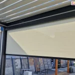 Widok z wnętrza nowoczesnej pergoli tarasowej SB400 na system bocznych osłon przeciwsłonecznych typu screen. Na pierwszym planie widoczny jest refleksol Ziiip 120 Aluprof w kolorze beżowym, który rozwinięty jest na część wysokości, stanowiąc skuteczną barierę przed słońcem i wiatrem. Tkanina charakteryzuje się gładkim splotem i jest stabilnie zamontowana w antracytowej ramie konstrukcyjnej pergoli. Powyżej rolety widoczne jest zadaszenie pergoli wykonane z ruchomych, aluminiowych lameli (system bioklimatyczny) w kolorze białym, ustawionych pod kątem, co pozwala na regulację dopływu światła. W tle, za konstrukcją pergoli, znajduje się ekspozycja zewnętrzna z różnymi rodzajami kostki brukowej, płyt tarasowych oraz elementami małej architektury ogrodowej. Całość prezentuje funkcjonalne wykorzystanie rolet screen Aluprof jako integralnego elementu nowoczesnych systemów zadaszeń tarasowych.