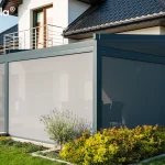 Nowoczesna pergola tarasowa o konstrukcji z ciemnych, grafitowych profili aluminiowych zamontowana przy nowoczesnym budynku mieszkalnym z jasną elewacją i ciemnym dachem skośnym. Zadaszenie wyposażone jest w boczne oraz frontowe rolety typu screen w kolorze szarym, które są całkowicie opuszczone, tworząc pełną osłonę przeciwsłoneczną i osłonę przed wiatrem dla przestrzeni pod pergolą. Konstrukcja przylega do ściany domu bezpośrednio pod balkonem z metalową balustradą ze stali nierdzewnej i przeszklonymi drzwiami balkonowymi. Wokół pergoli znajduje się starannie zaprojektowany ogród z trawnikiem, rabatami kwiatowymi oraz niskimi krzewami ozdobnymi, a całość prezentuje się jako eleganckie i funkcjonalne rozwiązanie zacieniające.