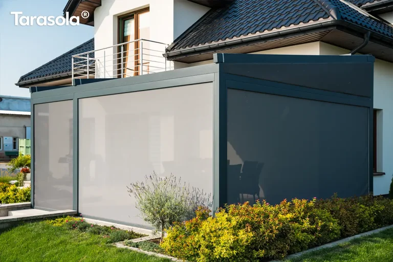 Nowoczesna pergola tarasowa o konstrukcji z ciemnych, grafitowych profili aluminiowych zamontowana przy nowoczesnym budynku mieszkalnym z jasną elewacją i ciemnym dachem skośnym. Zadaszenie wyposażone jest w boczne oraz frontowe rolety typu screen w kolorze szarym, które są całkowicie opuszczone, tworząc pełną osłonę przeciwsłoneczną i osłonę przed wiatrem dla przestrzeni pod pergolą. Konstrukcja przylega do ściany domu bezpośrednio pod balkonem z metalową balustradą ze stali nierdzewnej i przeszklonymi drzwiami balkonowymi. Wokół pergoli znajduje się starannie zaprojektowany ogród z trawnikiem, rabatami kwiatowymi oraz niskimi krzewami ozdobnymi, a całość prezentuje się jako eleganckie i funkcjonalne rozwiązanie zacieniające.