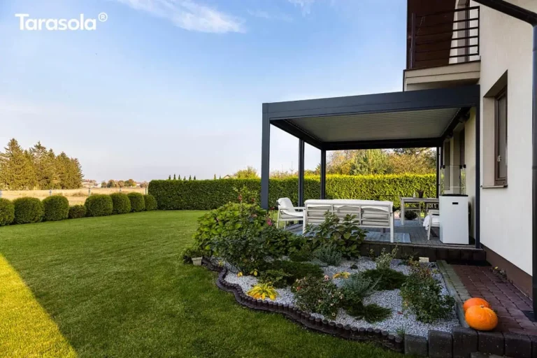 Zdjęcie przedstawia widok z boku na dom jednorodzinny z przylegającą do niego nowoczesną konstrukcją Essential tarasola w kolorze antracytowym. Pergola jest zamontowana do jasnej elewacji budynku, pod balkonem o prostej, metalowej balustradzie. Pod zadaszeniem z białych lameli znajduje się umeblowany taras z białą sofą i stolikiem kawowym, ustawionymi na geometrycznym, niebiesko-białym dywanie. Wokół tarasu rozciąga się starannie zaprojektowany ogród. Na pierwszym planie widoczna jest ozdobna rabata wysypana jasnym żwirem, na której rosną niskie krzewy iglaste, wrzosy i inne rośliny ozdobne, oddzielona od trawnika palisadą o falistym kształcie. Obok rabaty, na ścieżce z kostki brukowej, leżą dwie dekoracyjne pomarańczowe dynie. Dalszy plan stanowi duży, zielony trawnik, niski żywopłot z formowanych krzewów oraz wysoki, gęsty żywopłot odgradzający posesję. W oddali widać otwartą przestrzeń pól i pojedyncze drzewa pod bezchmurnym, błękitnym niebem.