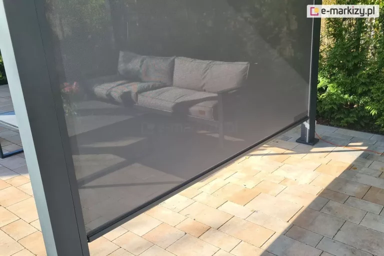 Zdjęcie prezentuje niemal całkowicie rozwinięty refleksol Zip z tkaniną typu screen, zamontowany w konstrukcji nowoczesnej pergoli tarasowej. Tkanina techniczna w kolorze ciemnego grafitu posiada drobny, siatkowy splot, który zapewnia prywatność i ochronę przeciwsłoneczną, zachowując jednocześnie częściową przezierność. Przez rozwiniętą przesłonę widać zarys sofy ogrodowej z szarymi poduszkami stojącej wewnątrz zacienionej strefy wypoczynkowej. Roleta porusza się w bocznej prowadnicy zintegrowanej z pionowym, masywnym słupem pergoli o matowym, antracytowym wykończeniu. Dolna krawędź materiału jest usztywniona poziomą listwą obciążającą w tym samym kolorze co stelaż. Pergola posadowiona jest na tarasie wyłożonym prostokątną kostką brukową w ciepłych, beżowo-piaskowych odcieniach. W tle, po prawej stronie, widoczny jest gęsty, zielony żywopłot z tui, a po powierzchni tarasu biegnie pomarańczowy przewód elektryczny. Mocne, boczne światło słoneczne podkreśla strukturę tkaniny i precyzję montażu systemu osłonowego.