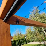 Pergola Koro z dachem Supro mocowanym pod belką z widocznym elementem napędu ręcznego sznurkowego. W tle ogród z trawnikiem i miejscem wyłożonym kostką oraz placem zabaw dla dzieci. W tle drzewa, żywopłot i błękitne niebo.