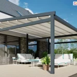 Zdjęcie prezentuje wizualizację nowoczesnej pergoli tarasowej Moderno wolnostojącej, osadzonej na rozległym tarasie przy budynku o współczesnej architekturze. Konstrukcja wykonana jest z profili aluminiowych w kolorze antracytowym, wsparta na masywnych słupach podporowych. Zadaszenie stanowi jasnokremowa, segmentowa tkanina, która jest całkowicie rozłożona i napięta na poprzecznych profilach, chroniąc przestrzeń przed słońcem. Pod pergolą znajduje się luksusowa strefa wypoczynkowa z białymi sofami modułowymi o turkusowych akcentach, uzupełniona białymi stolikami kawowymi i dekoracyjnymi lampionami. Elewacja domu łączy biały tynk z ciemną cegłą klinkierową oraz dużymi, panoramicznymi przeszkleniami. W tle widoczny jest zielony ogród z drzewami pod bezchmurnym, niebieskim niebem. Scena podkreśla wysoką estetykę oraz funkcjonalność zautomatyzowanego systemu zadaszenia dużych powierzchni tarasowych.