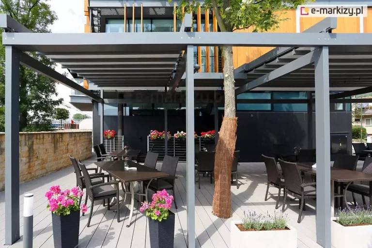 Nowoczesny ogródek restauracyjny osłonięty wielomodułową pergolą tarasową Solid Aluprof o konstrukcji aluminiowej w kolorze antracytowym. Zadaszenie składa się z szarego, segmentowego materiału. Pod pergolą, na jasnym tarasie z desek kompozytowych, ustawiono ciemne stoły i technorattanowe fotele. Konstrukcja została wkomponowana w otoczenie, uwzględniając rosnące drzewo liściaste, którego pień owinięto brązową matą ochronną. Przestrzeń udekorowana jest licznymi kwiatami: różowymi pelargoniami w wysokich antracytowych donicach oraz lawendą w białych, prostokątnych skrzyniach. W tle widoczny jest nowoczesny budynek z elewacją łączącą czarne panele z pionowymi drewnianymi lamelami oraz kamienny murek po lewej stronie. Całość oświetlona jest naturalnym, dziennym światłem.