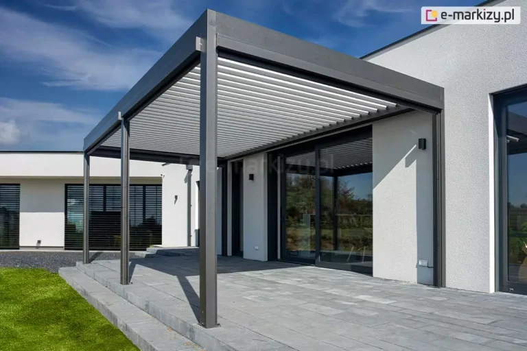 Nowoczesna pergola bioklimatyczna przyścienna w kolorze antracytowym, zamontowana na podwyższonym tarasie z szarych płyt betonowych. Konstrukcja zadaszenia składa się z białych, aluminiowych lameli ustawionych w pozycji uchylonej. Pergola jest wsparta na solidnych, kwadratowych słupach i przylega do białej, tynkowanej elewacji nowoczesnego domu o parterowej bryle. W elewacji widoczne są duże, przeszkolone drzwi tarasowe w ciemnych ramach oraz czarne, minimalistyczne kinkiety zewnętrzne. W tle znajduje się dalsza część budynku z oknami wyposażonymi w czarne żaluzje fasadowe oraz fragment zielonego trawnika i niebieskie niebo z lekkimi chmurami. Zdjęcie prezentuje minimalistyczną architekturę i integrację systemu zadaszenia z bryłą budynku.