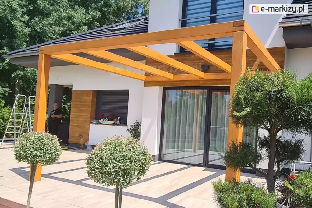 Drewniana przyścienna pergola zamocowana nad drzwiami tarasowymi do elewacji budynku o jasnej fasadzie. Przed tarasem widoczne drzewko iglaste i dwa drzewka liściaste o kulistej koronie. W tle na tarasie stoi mężczyzna, obok którego ustawiono dwie drabiny.