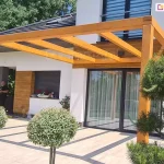 Drewniana przyścienna pergola zamocowana nad drzwiami tarasowymi do elewacji budynku o jasnej fasadzie. Przed tarasem widoczne drzewko iglaste i dwa drzewka liściaste o kulistej koronie. W tle na tarasie stoi mężczyzna, obok którego ustawiono dwie drabiny.