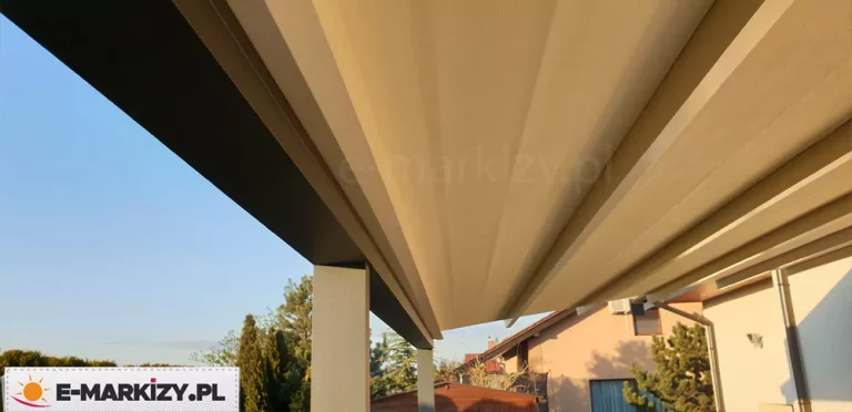 Zdjęcie prezentuje ujęcie od dołu nowoczesnej pergoli rzymskiej Luxo, ukazujące strukturę rozłożonego poszycia. Zadaszenie wykonane jest z wysokiej jakości tkaniny w kolorze piaskowym lub jasnobeżowym, która układa się w estetyczne, równoległe pasy dzięki poprzecznym profilom aluminiowym. Konstrukcja nośna, widoczna po lewej stronie, posiada matowe wykończenie w kolorze ciemnego antracytu i opiera się na solidnych słupach o przekroju kwadratowym. W tle, pod zadaszeniem, widoczny jest fragment ogrodu z drzewami iglastymi oraz sąsiednie budynki mieszkalne o jasnych elewacjach na tle bezchmurnego, błękitnego nieba. Perspektywa podkreśla precyzję wykonania mechanizmu tkaninowego oraz minimalistyczny design osłony tarasowej, która skutecznie chroni przestrzeń wypoczynkową przed intensywnym nasłonecznieniem.
