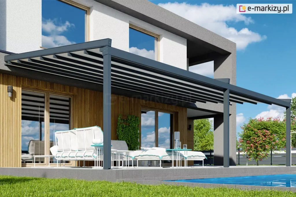 Nowoczesna pergola tarasowa Sqaure o konstrukcji aluminiowej w kolorze ciemnoszarym zamontowana przy elewacji piętrowego budynku mieszkalnego. Zadaszenie pergoli składa się z ruchomego poszycia z tkaniny w kolorze białym, które jest rozsunięte, dzięki czemu widoczne są ciemne profile przeciwwietrzne. Konstrukcja opiera się na trzech solidnych pionowych słupach o przekroju kwadratowym, które są zakotwiczone w podłożu tarasu wykonanego z szarych płyt. Budynek posiada elewację łączącą biały tynk na piętrze z drewnianą okładziną z pionowych lameli na parterze oraz ciemnoszare elementy konstrukcyjne wnęki tarasowej. Pod pergolą znajduje się biały zestaw mebli ogrodowych z jasnymi siedziskami wykończonymi błękitną lamówką, a także dwa białe stoliki kawowe o ażurowej podstawie. W tle widoczny jest fragment basenu z błękitną wodą, zielony trawnik, niskie nasadzenia roślinne oraz krzew o różowych kwiatach. Całość kompozycji przedstawiona jest w pełnym słońcu przy bezchmurnym, niebieskim niebie, które odbija się w dużych przeszkleniach okiennych budynku.