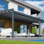 Nowoczesna pergola tarasowa Sqaure o konstrukcji aluminiowej w kolorze ciemnoszarym zamontowana przy elewacji piętrowego budynku mieszkalnego. Zadaszenie pergoli składa się z ruchomego poszycia z tkaniny w kolorze białym, które jest rozsunięte, dzięki czemu widoczne są ciemne profile przeciwwietrzne. Konstrukcja opiera się na trzech solidnych pionowych słupach o przekroju kwadratowym, które są zakotwiczone w podłożu tarasu wykonanego z szarych płyt. Budynek posiada elewację łączącą biały tynk na piętrze z drewnianą okładziną z pionowych lameli na parterze oraz ciemnoszare elementy konstrukcyjne wnęki tarasowej. Pod pergolą znajduje się biały zestaw mebli ogrodowych z jasnymi siedziskami wykończonymi błękitną lamówką, a także dwa białe stoliki kawowe o ażurowej podstawie. W tle widoczny jest fragment basenu z błękitną wodą, zielony trawnik, niskie nasadzenia roślinne oraz krzew o różowych kwiatach. Całość kompozycji przedstawiona jest w pełnym słońcu przy bezchmurnym, niebieskim niebie, które odbija się w dużych przeszkleniach okiennych budynku.