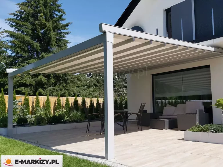 Nowoczesna pergola tarasowa o konstrukcji aluminiowej w kolorze jasnoszarym lub srebrnym, zamontowana przy białej elewacji nowoczesnego budynku mieszkalnego. Zadaszenie pergoli wykonane jest z jasnego, kremowego materiału ułożonego w charakterystyczne, poziome pasy, wsparte na systemie poprzecznych profili. Konstrukcja opiera się na dwóch solidnych, kwadratowych słupach bocznych, a jej tylna część jest przytwierdzona bezpośrednio do ściany domu powyżej dużego przeszklenia tarasowego. Pod pergolą znajduje się strefa wypoczynkowa wyposażona w ciemnoszare meble technorattanowe z jasnymi poduszkami oraz zestaw mebli ogrodowych składający się ze stołu i krzeseł o metalowej ramie. Taras wyłożony jest jasnymi płytami o strukturze drewna. Wzdłuż krawędzi tarasu przebiega niska, biała donica z nasadzeniami roślinnymi, a w tle widoczne jest drewniane ogrodzenie, za którym rośnie szpaler wysokich tui oraz duże drzewo iglaste. Na piętrze budynku widoczne są nowoczesne barierki balkonowe i ciemne rolety zewnętrzne. Całość sceny skąpana jest w jasnym, dziennym świetle przy bezchmurnym, błękitnym niebie.