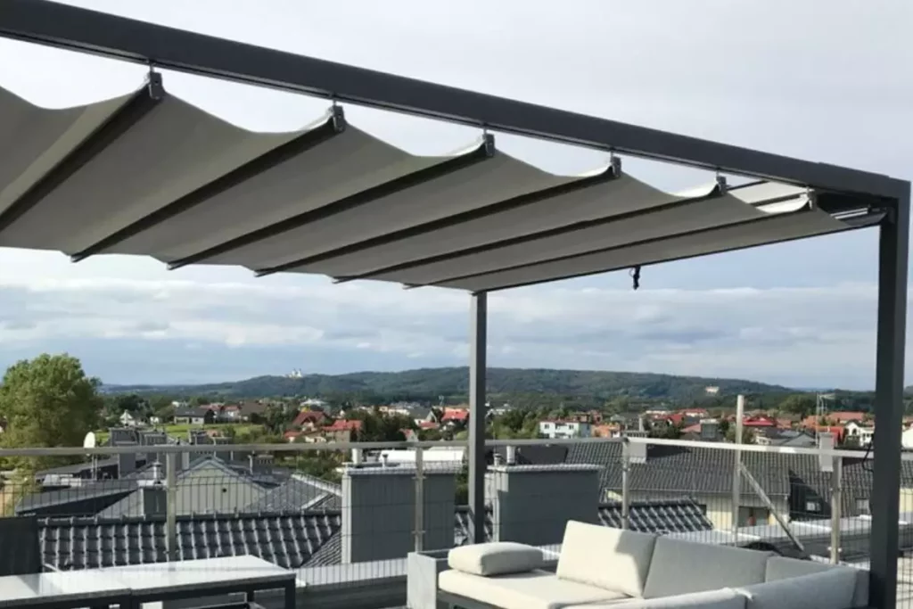 Widok z bliska na system zadaszenia nowoczesnej pergoli wolnostojącej o antracytowej, aluminiowej konstrukcji, zainstalowanej na tarasie dachowym. Zdjęcie koncentruje się na detalach jasnej, technicznej tkaniny, która jest częściowo rozwinięta i tworzy charakterystyczne, miękkie fale podwieszone pod poprzecznymi profilami prowadzącymi. Materiał jest zamocowany do czarnych prowadnic za pomocą systemowych uchwytów, co pozwala na jego swobodne przesuwanie wzdłuż ramy. W tle rozpościera się szeroka panorama podmiejskiego osiedla z domami jednorodzinnymi o ciemnych dachach, widoczna ponad metalową barierką zabezpieczającą taras. Niebo jest lekko zachmurzone, w odcieniach jasnego błękitu i szarości. W dolnej części kadru, pod zadaszeniem, widoczny jest fragment jasnoszarej sofy ogrodowej z miękkimi poduchami oraz krawędź ciemnego stolika kawowego.