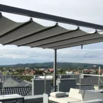 Widok z bliska na system zadaszenia nowoczesnej pergoli wolnostojącej o antracytowej, aluminiowej konstrukcji, zainstalowanej na tarasie dachowym. Zdjęcie koncentruje się na detalach jasnej, technicznej tkaniny, która jest częściowo rozwinięta i tworzy charakterystyczne, miękkie fale podwieszone pod poprzecznymi profilami prowadzącymi. Materiał jest zamocowany do czarnych prowadnic za pomocą systemowych uchwytów, co pozwala na jego swobodne przesuwanie wzdłuż ramy. W tle rozpościera się szeroka panorama podmiejskiego osiedla z domami jednorodzinnymi o ciemnych dachach, widoczna ponad metalową barierką zabezpieczającą taras. Niebo jest lekko zachmurzone, w odcieniach jasnego błękitu i szarości. W dolnej części kadru, pod zadaszeniem, widoczny jest fragment jasnoszarej sofy ogrodowej z miękkimi poduchami oraz krawędź ciemnego stolika kawowego.