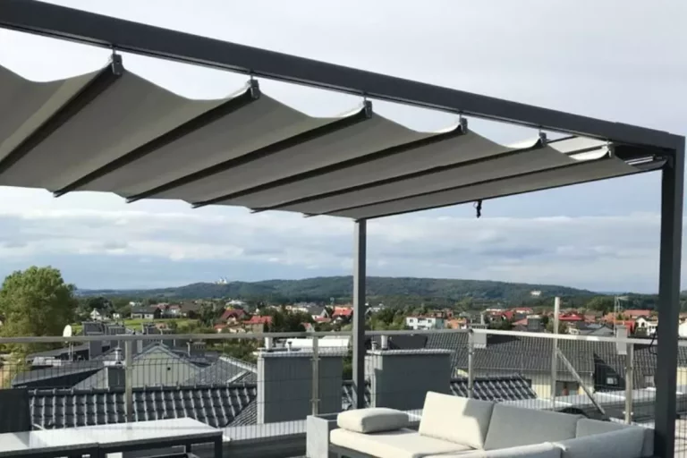 Widok z bliska na system zadaszenia nowoczesnej pergoli wolnostojącej o antracytowej, aluminiowej konstrukcji, zainstalowanej na tarasie dachowym. Zdjęcie koncentruje się na detalach jasnej, technicznej tkaniny, która jest częściowo rozwinięta i tworzy charakterystyczne, miękkie fale podwieszone pod poprzecznymi profilami prowadzącymi. Materiał jest zamocowany do czarnych prowadnic za pomocą systemowych uchwytów, co pozwala na jego swobodne przesuwanie wzdłuż ramy. W tle rozpościera się szeroka panorama podmiejskiego osiedla z domami jednorodzinnymi o ciemnych dachach, widoczna ponad metalową barierką zabezpieczającą taras. Niebo jest lekko zachmurzone, w odcieniach jasnego błękitu i szarości. W dolnej części kadru, pod zadaszeniem, widoczny jest fragment jasnoszarej sofy ogrodowej z miękkimi poduchami oraz krawędź ciemnego stolika kawowego.